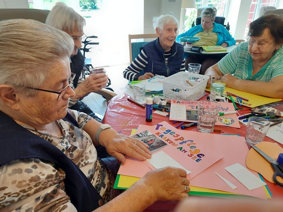 Bild zeigt vier Besucherinnen der Tagespflege an einem Tisch sitzend und Karten und Bilder gestaltend. Es werden Fotos auf A4 Blätter (buntes Tonpapier) geklebt und mit Stiften in bunten Buchstaben der Name der Tagespflege geschrieben. Die Besucherinnen der Tagespflege haben sichtlich Spaß daran.