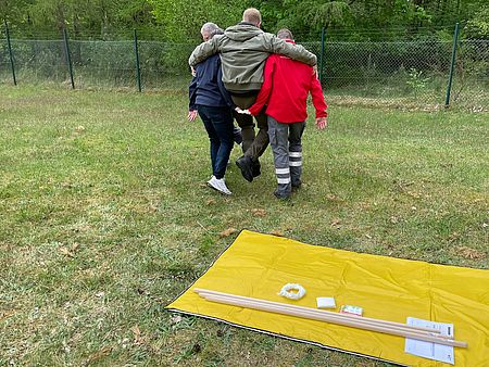 Bild zeigt, wie eine Person (Verletzter) mit dem Zwei-Hände-Griff von zwei Helfern transportiert wird