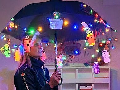 Bild zeigt eine junge Frau mit einem aufgespannten Regenschirm an dem eine batteriebetriebene Lichterkette und verschiede Kleinspielteile die auch beleuchtet sind, angeberacht.