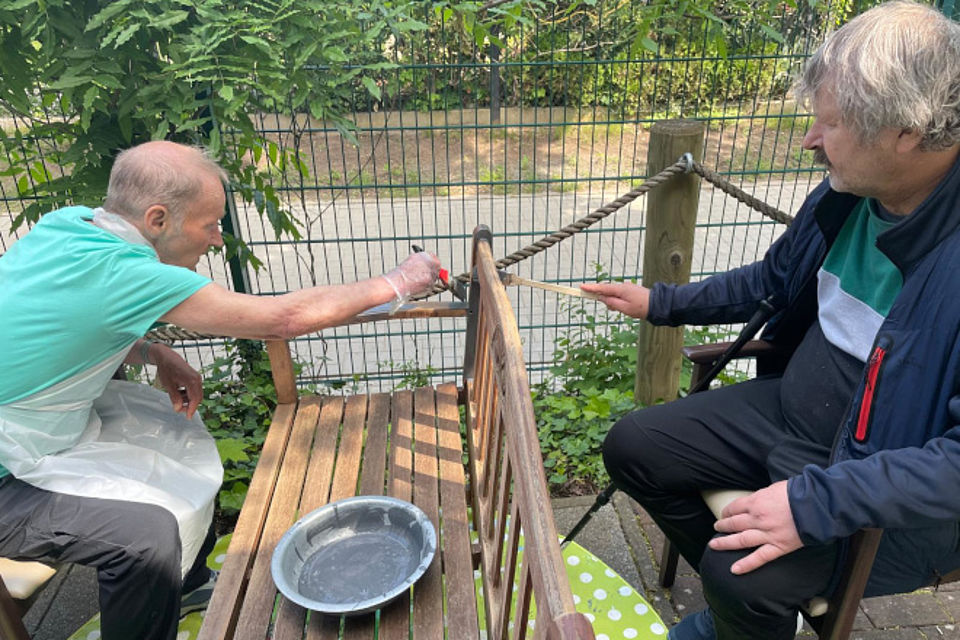 Bild zeigt: Zwei Männer sind im Freien mit Handwerksarbeiten beschäftigt. Ein älterer Mann (links) in einem mintgrünen Shirt und mit einer Plastikschürze streicht eine hölzerne Gartenbank mit einem Pinsel. Der andere Mann (rechts) sitzt ihm gegenüber, trägt eine dunkle Jacke und streicht ebenfalls mit einem Pinsel. In der Mitte steht eine Schale mit der Farbe. Im Hintergrund sind ein Zaun und ein Seilgeländer sichtbar. Die Szene zeigt eine gemeinsame handwerkliche Aktivität und Gartenpflege in der Tagespflege.