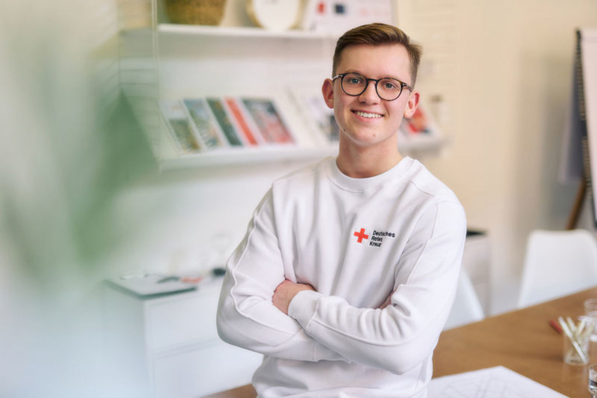 Bild zeigt einen jungen Mann mit vor der Brust verschränkten Armen auf einem Tisch sitzend. Er trägt ein weißes Sweatshirt mit einem Rotkreuz Logo.