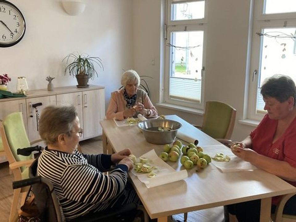 Bild zeigt zwei Besucherinnen der Tagespflege mit einer Pflegefachkraft an einem Tisch im Gemeinschaftsraum sitzend und Äpfel schälen. Auf dem Tisch liegen noch ca. 15 Äpfel und es steht eine große Edelstahlschale in der Mitte, in der die geschälten Äpfel liegen.