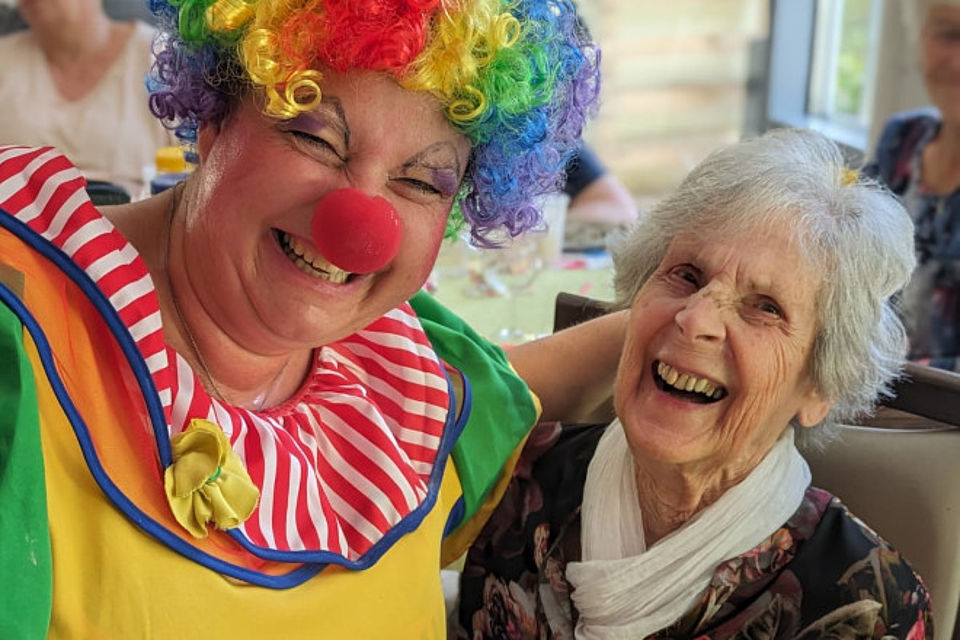 Bild zeigt: Nahaufnahme von zwei lachenden Frauen beim Zirkusfest in der Tagespflege. Links ist eine Frau als Clown verkleidet, mit einem roten Ringelkragen, gelb-rotem Oberteil, roter Nase und einer bunten Regenbogenperücke. Die Clownin umarmt freundlich eine ältere Dame (rechts) mit weißen Haaren, die ebenfalls herzlich lacht und ein weißes Halstuch trägt. Die Szene vermittelt Freude und gute Stimmung.