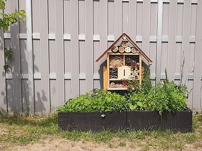 Bild zeigt ein recht großes Insektenhotel mit davor angelegtem Wildpflanzenbewuchs in Hochbeet ähnlichen Kästen. Das Insektenhotel steht vor einer grauen Holzzaunwand.