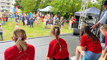 Bild zeigt einen Blick von der Bühne auf das Festgelände mit ganz vielen Besuchern, Erwachsenen und Kindern. Auf der Bühne sind die Kinder der Trommelgruppe "Hort Stelzenvilla" in einer Rückenansicht zu sehen.