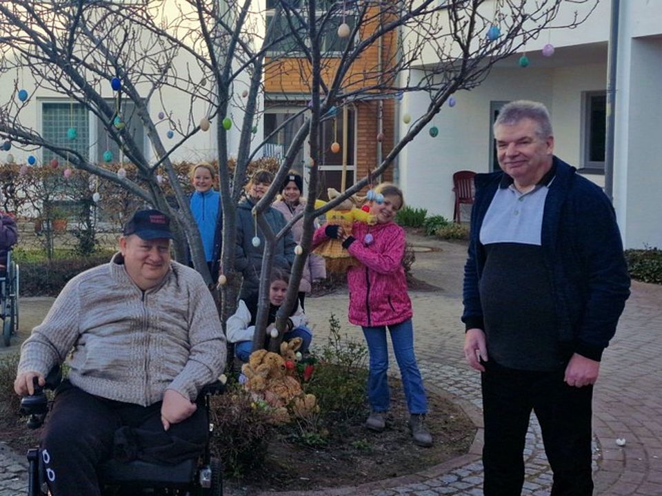 Bild 4 Bild zeigt einen Gast der TAgespflege im Rollstuhl im Außengelände vor einem Buschähnlichem Baum. Dieser ist an den Zweigen mit den bemalten Eiern behängt. Im Hintergrund stehen bzw. hocken 5 Kinder. Rechts neben dem Baum steht noch ein weiterer Tagesgast.