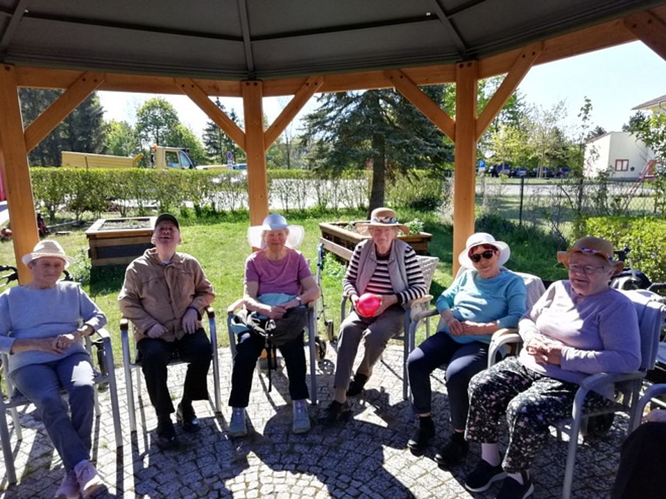 Bild zeigt eine Gruppe (6) von Besuchern der Tagespflege im Außenbereich bei sonnigem Wetter unter dem Pavillon im Kreis sitzend. Eine Besucherin hält einen roten Ball in ihren Händen, der dann im Kreis weitergegeben wird.