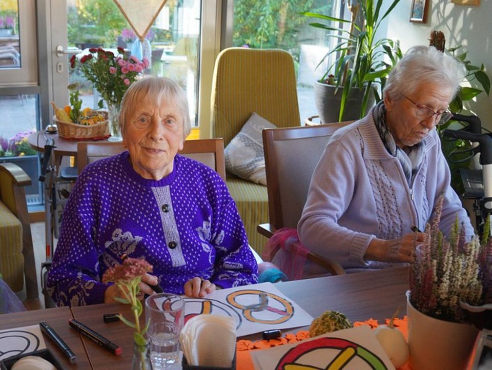 Bild zeigt zwei Besucherinnen der Tagespflege an einem Tisch sitzend. Vor ihnen liegt je ein Blatt Papier mit großen aufgedruckten Brezeln, welche mit farbigen Stiften in verschiedenen Farben ausgemalt werden. Im Vordergrund sieht man herbstliche Blumen auf dem Tisch stehen, sowie im Hintergrund auf einem Tisch ein Korb mit verschiedenen Zierkürbissen und eine Vase mit einem Straß Herbstastern.
