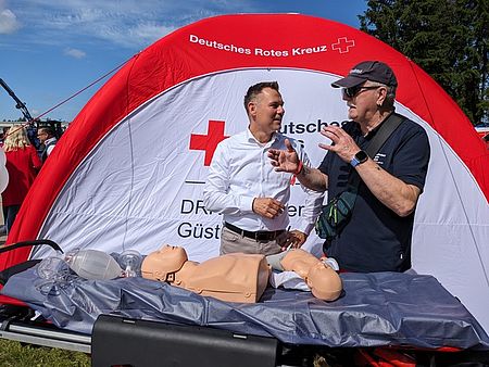 Bild zeigt Güstrows Bürgermeister Sascha Zimmermann hinter einer Rettungstrage stehend im Gespräch mit Wolf-Ulrich Pettke. Auf der Trage liegt eine Übungspuppe für Wiederbelebeungsmaßnahmen. Im Hintergrund ist ein Rot-Kreuz-Zelt zu sehen