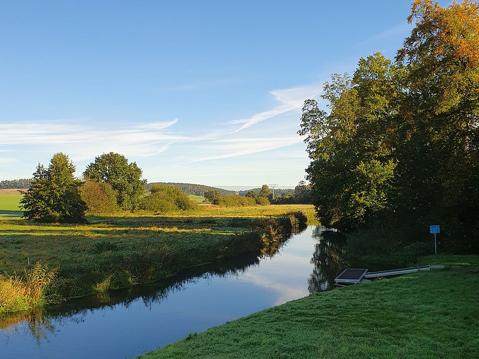 Bild zeigt: Eine ruhige Landschaftsaufnahme an einem sonnigen Tag mit klarem blauem Himmel. Ein schmaler, dunkelblauer Fluss, der leicht nach links abbiegt, spiegelt den Himmel und die Ufervegetation wider. Die Uferböschung im Vordergrund ist mit taunassem, hellem Gras bewachsen. Rechts und im Hintergrund säumen dichte grüne und leicht gelbliche Bäume das Ufer. Ein kleiner Steg ist auf der rechten Seite des Flusses zu erkennen.