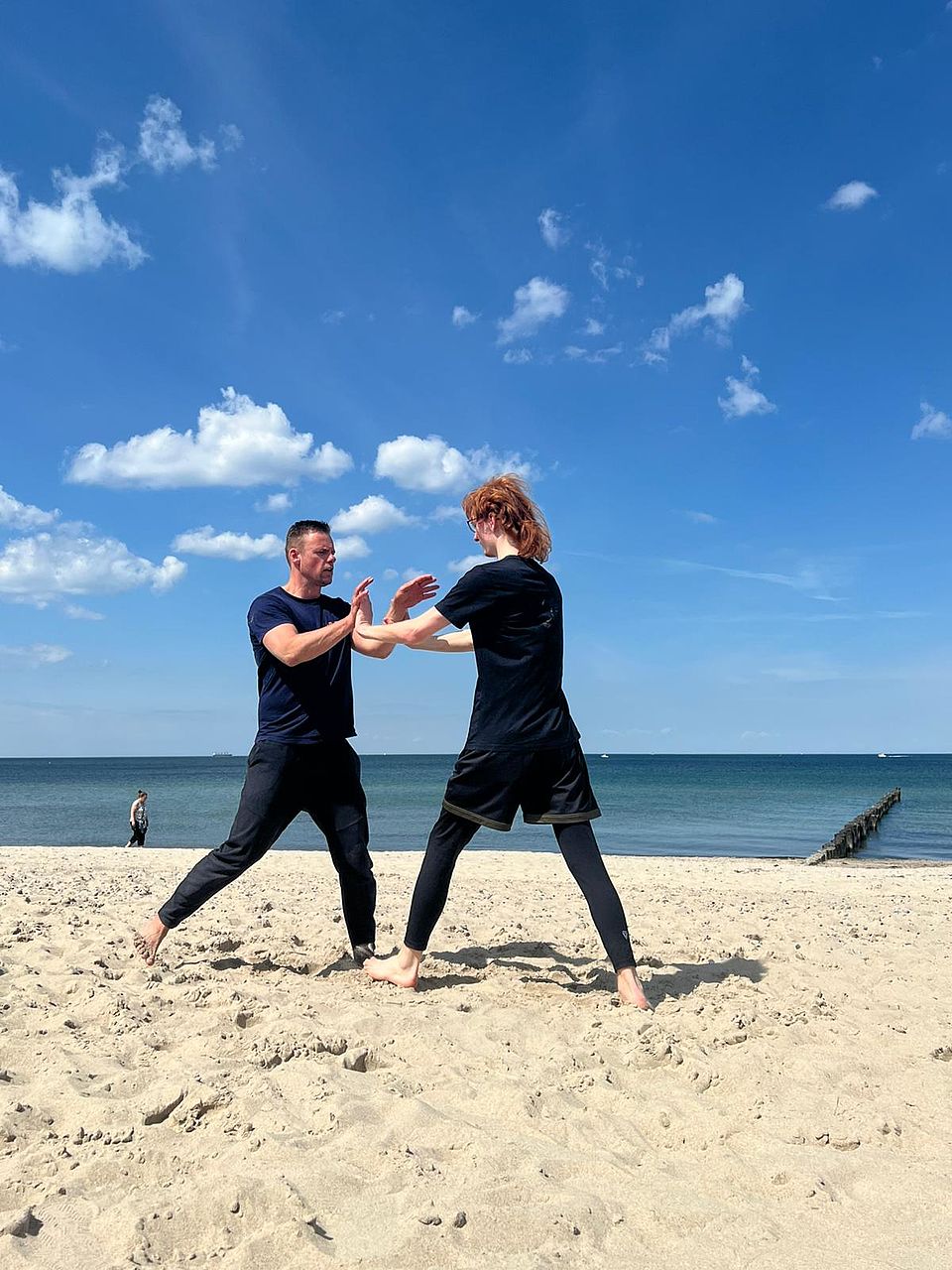 Bild zeigt eine junge Frau mit dem Übungsleiter am Strand der Ostsee sich gegenüber stehend. Sie haben beide eine Kampfhaltung und die Frau versucht einen Angriff abzuwehren.