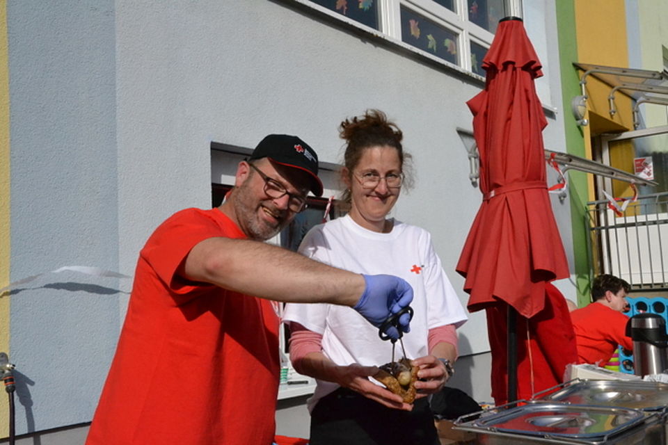 Bild zeigt Mitarbeitende (ein Mann und eine Frau) des Ernährungszentrums am Grillstand. Die Frau hält ein aufgeschnittenes Körnerbrötchen in der Hand und der Mann legt mit einer Grillzange eine Bratwurst in das Brötchen. Beide lachen freundlich in die Kamera. Im Hintergrund ist das Gebäude der Kita zu sehen.