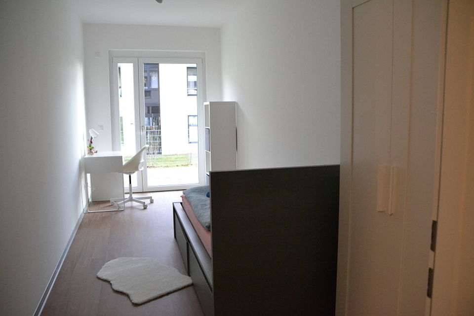 Bild zeigt den Blick in ein Einzelzimmer von der Tür aus, mit Schrank, Bett, Regal und Schreibtisch