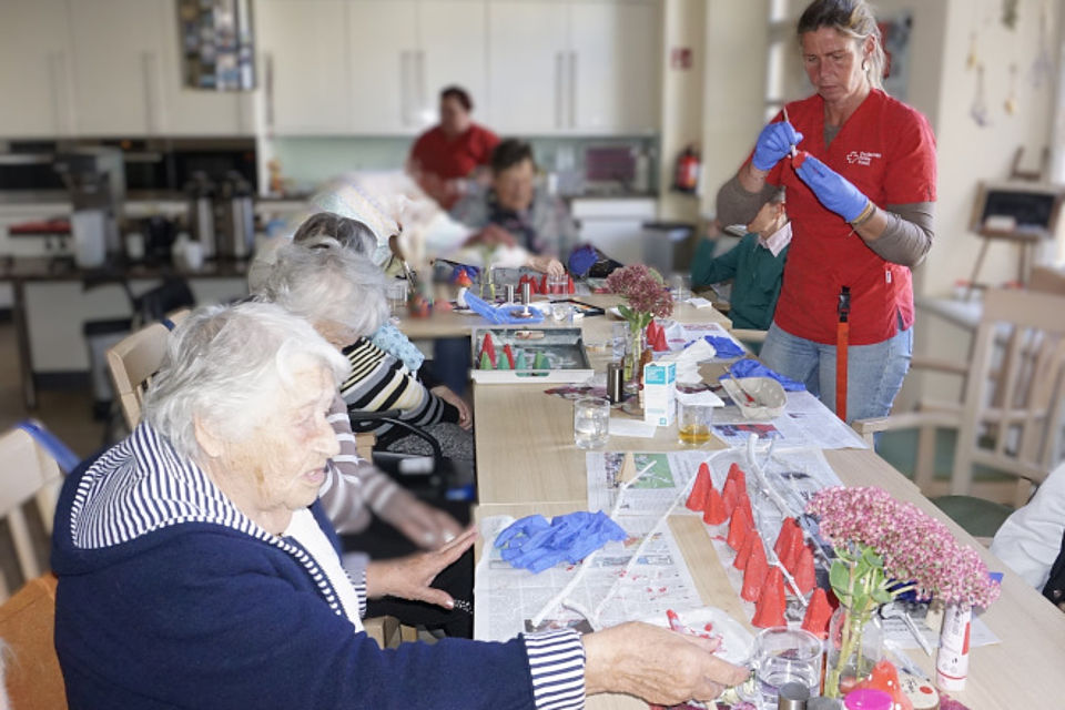 Bild zeigt: Eine Gruppe von Senioren nimmt an einer kreativen Bastelstunde in einem Gemeinschaftsraum teil. Eine Mitarbeiterin (rechts) in roter Dienstkleidung und blauen Handschuhen ist dabei, kleine rote Dekorationsobjekte zu bekleben oder zu bemalen. Die Senioren sitzen an einem langen Tisch, auf dem Bastelmaterialien, Handschuhe und Zeitungen zum Schutz ausgebreitet sind. Die ältere Dame im Vordergrund (links) in einem dunkelblauen Oberteil greift nach einem Material. Die Szene zeigt eine Aktivierungsgruppe beim gemeinsamen Basteln in der Tagespflege.