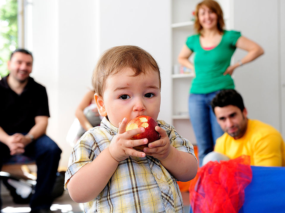 Bild zeigt mehrere Erwachsene Personen, Frauen und Männer, die bei einem Kurs Eltern und Kinder teilnehmen. im Vordergrund ist ein kleiner Junge zu sehen, welcher einen Apfel isst.