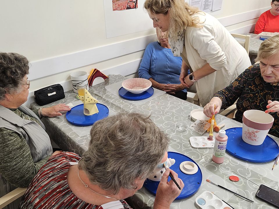 Bild zeigt: Mehrere Seniorinnen sitzen in einem hellen Raum an einem Tisch und bemalen Keramikgefäße. Eine junge Frau in einem hellen Pullover beugt sich über eine ältere Frau, um ihr Anweisungen zu geben. Vor den Teilnehmerinnen stehen verschiedene Keramikrohlinge, wie eine Schale und Blumentöpfe, sowie kleine Farbtöpfe, Pinsel und Schwämme. Eine Seniorin im Vordergrund ist gerade dabei, eine Figur mit einem kleinen Pinsel zu bemalen.