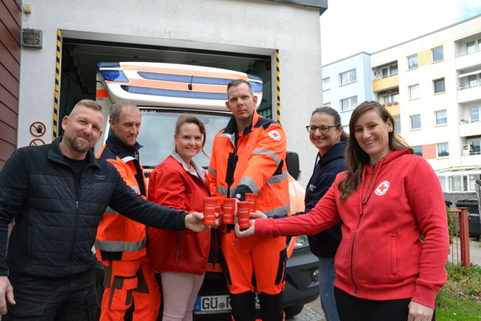 Bild zeigt 6 DRK Mitarbeitende, teils in leitender Position, die zusammen stehen und die Rot Kreuz Dose in die Mitte halten. Im Hintergrund ist ein DRK Rettungsfahrzeug zu sehen.