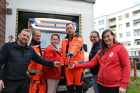 Bild zeigt 6 DRK Mitarbeitende, teils in leitender Position, die zusammen stehen und die Rot Kreuz Dose in die Mitte halten. Im Hintergrund ist ein DRK Rettungsfahrzeug zu sehen.