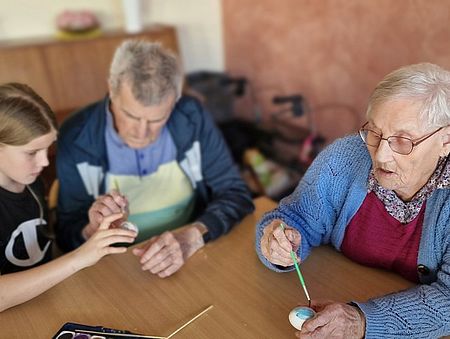 Bild zeigt ein Mädchen sowie einen Senior und eine Seniorin am Tisch sitzend, die zusammen ausgeblasenen Eier mit Wasserfarben bemalen.