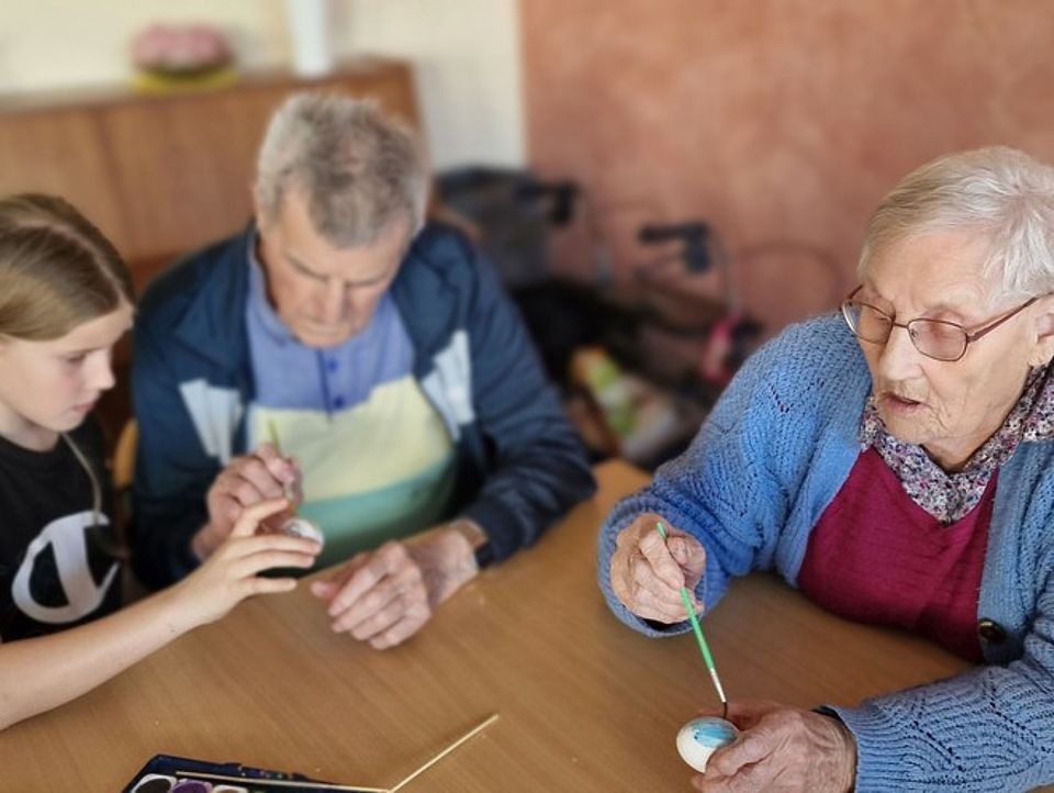 Bild 2 Bild zeigt ein Mädchen sowie einen Senior und eine Seniorin am Tisch sitzend, die zusammen ausgeblasenen Eier mit Wasserfarben bemalen.