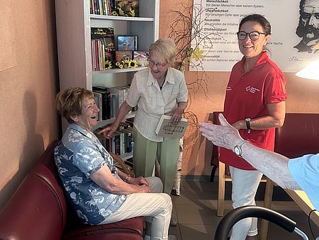 Bild zeigt zwei Bewohnerinnen des DRK Seniorenheim vor der neuen Büchertauschecke. Eine sitz auf einem dunkelroten Ledersofa, die andere Frau steht mit einem Buch in der Hand davor. Beide lachen fröhlich. Im Hintergrund ist das Regal mit den Tauschbüchern zu sehen. im Vordergrund steht eine Mitarbeiterin des Heimes.