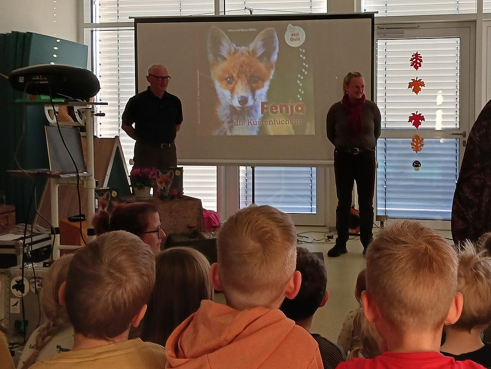 Vorlesewoche Bild 2 Bild zeigt Silva und Mario Müller vor einer Leinwand auf der das Bild von einem Fuchs zu sehen ist. Daneben ist das Wort "Fenja" zu lesen. Im Vordergrund sind Hinterköpfe von Kindern zu sehen und im seitlichen Profil das Gesicht einer Erzieherin.
