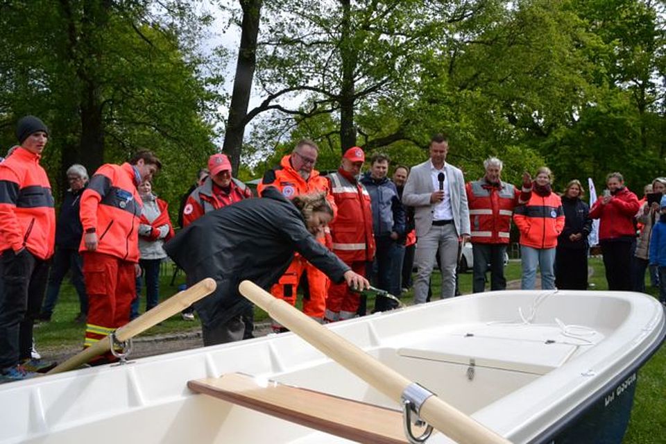 Bootstaufe Bild zeigt das neue Boot im Vordergrund. Dahinter stehen viele Besucher des Festes zum Teil in DRK Einsatzkleidung. Frau Mater (Stadträtin) steht gebückt am Bug des Bootes und gießt aus einer Sektflasche einen kleinen Schluck Sekt auf den Bug des Bootes.