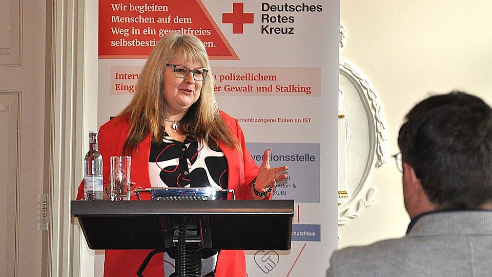 Bild zeigt Frau Jacqueline Garske bei ihrer Rede zum Trägerwechsel des Frauenschutzhauses. Sie steht hinter einem Rednerpult, auf der linken Seite steht eine Flasche Wasser mit einem Glas, im Hintergrund ist ein Roll-Up Aufsteller mit DRK Logo und Informationen zum Frauenschutzhaus