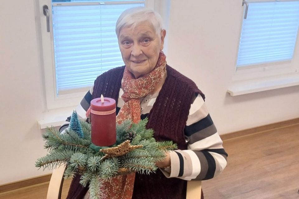 Bild zeigt eine Besucherin der Tagespflege auf einem Stuhlsessel sitzend. Sie hält ein Adventsgesteck mit einer dunkelroten LED-Kerze, die auf einem Bett aus Tannenzweigen steht.
