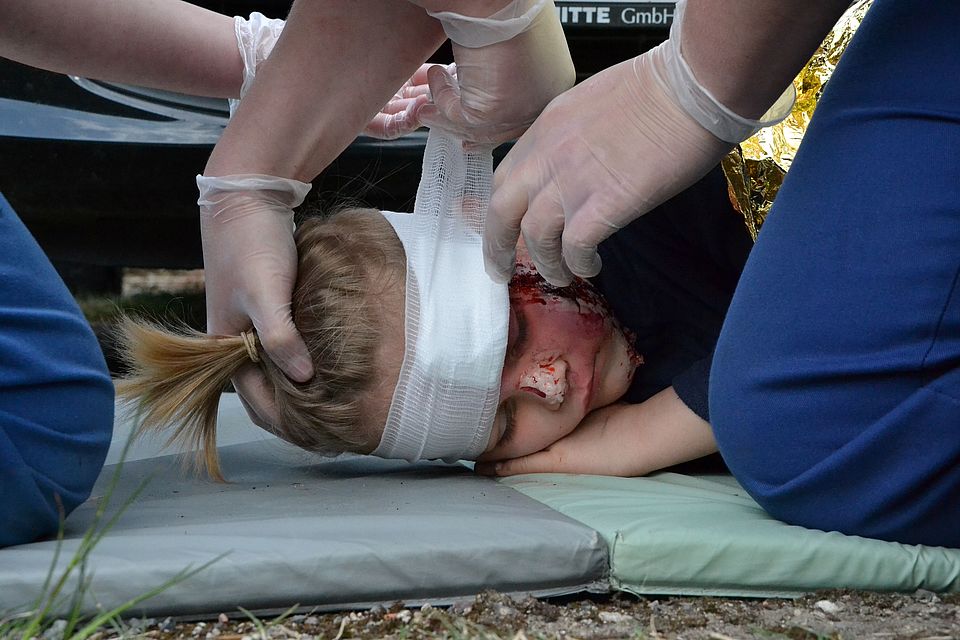 Bild zeigt in Nahaufnahme, wie bei einer Statistin (Mime) eine geschmickte Kopfverletzung versorgt wird. Vier Hände kümmern sich um die Verletzte und man sieht wie den Kopf halten und stützen, wärend die anderen beiden Hände einen Verband anlegen.