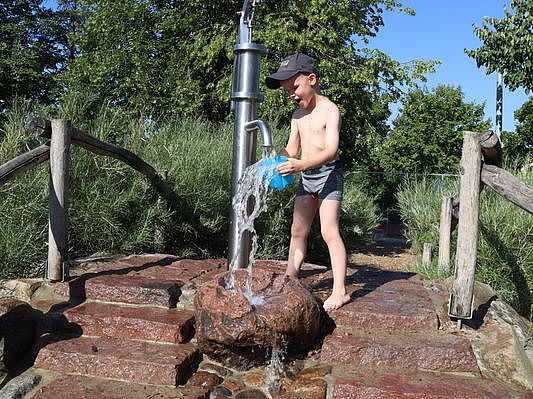 Bild zeigt einen Jungen an der Wasserpumpe vor der Sandspielanlage