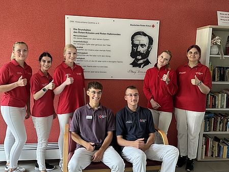 Bild zeigt 5 junge Frauen und zwei junge Männer. Die Frauen stehend in roten DRK Shirts und Jacken, die Männer sitzend in blauen DRK Polo-Shirts. Im Hintergrund  ist ein Wandbild mit dem Konterfei von Henry Dunant, Grunder der Rot Kreuz Bewegung, zu sehen und daneben die 7 Grundsätze des Roten Kreuzes