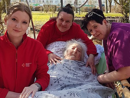 Bild zeigt Frau Wilma Eggert, liegend in ihrem Pflegebett. Das Bett wurde von den Mitarbeitenden Lara Holz und Daniela Blaubach, die beiden stehen links und am Kopfende des Bettes,  in die Grünanlage geschoben. Rechts am Bett steht die Tochter von Frau Eggert. Frau Eggert schaut zufrieden und glücklich in die Kamera.