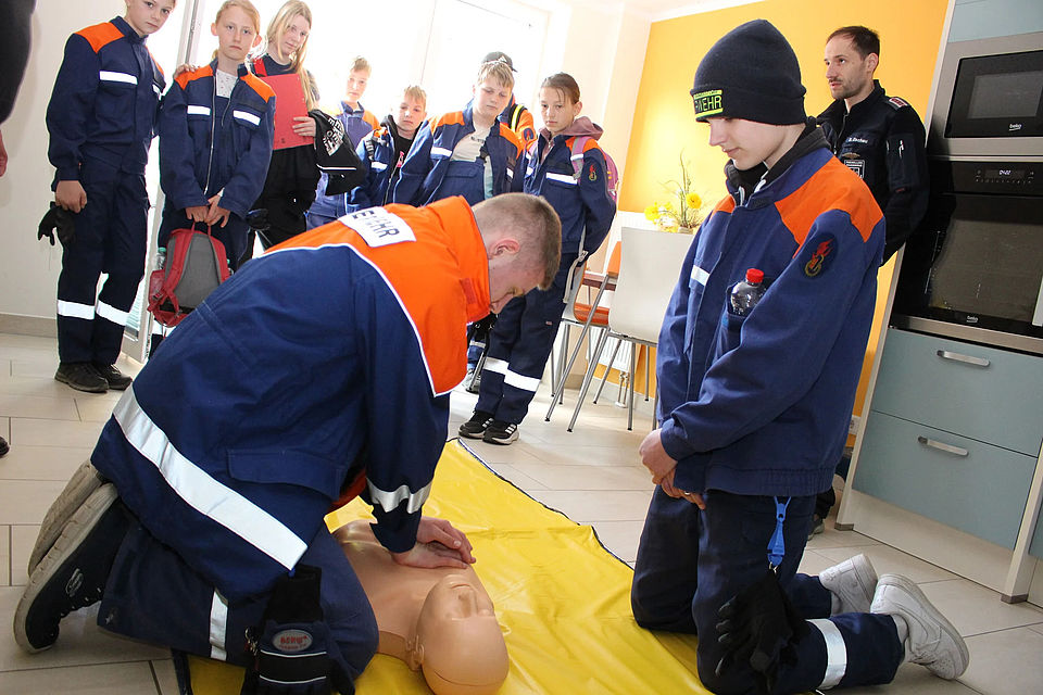 Bild zeigt eine Gruppe von Kindern der Feuerwehrgruppe. Ein Kind (Jugendlicher) beugt sich über eine Übungspuppe um an der Wiederbelebungsmaßnahmen (Herzdruckmassage und Beatmung) durchzuführen