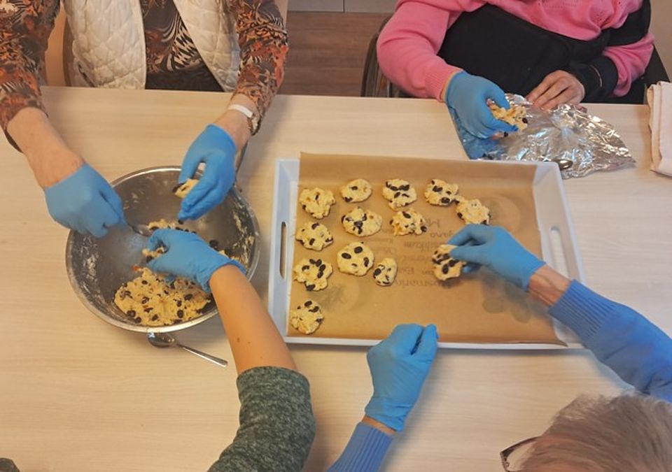 Bild zeigt vier paar Hände über einem Tisch auf dem eine Schüssel und ein Kucheblech stehen. Auf dem Blech liegen schon einige Weihnachtskekse und in der Schüssel ist noch ein wenig Teig zusehen.