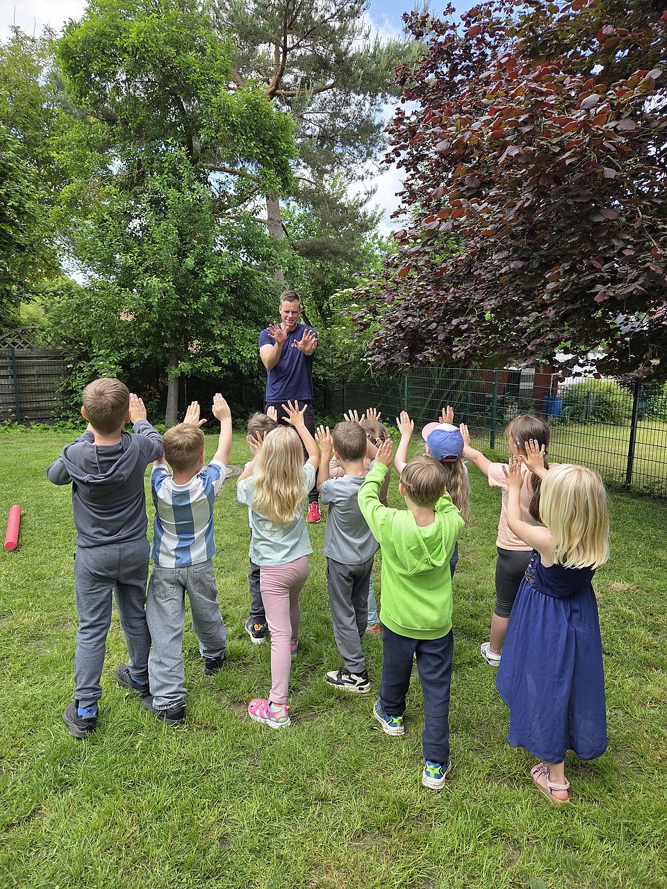 Bild zeigt eine Gruppe von kleinen Kindern, im Außengelände der Kita, stehen auf einer Wiese und wiederholen die Bewegungen des Übungsleiters