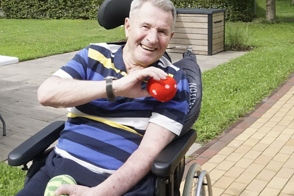 Bild zeigt: einen männlichen Tagesgast im Rollstuhl sitzend und an einer sportlichen Aktivität teilnehmend. Er hält in der rechten Hand einen einen roten Stoffball mit weißen Punkten und will diesen Ball gerade werfen. Er trägt ein blau-weiß-gelb gestreiftes Poloshirt. Der Rollstuhl steht auf einem gepflasterten Weg. Im Hintergrund sieht man ein wenig Rasenfläche und einen Teil einer Hecke.
