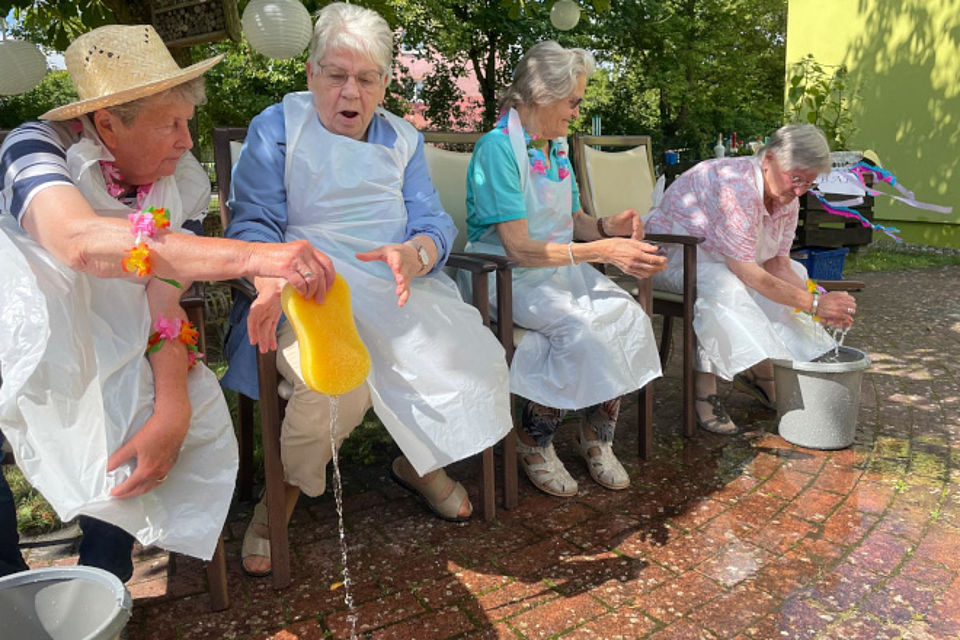 Bild zeigt: Vier ältere Damen nehmen lachend und begeistert an einem lustigen Wasserspiel im Freien teil. Alle sitzen auf Stühlen in einer Reihe, tragen weiße Schürzen zum Schutz vor Nässe und haben links und rechts der Reihe je einen Eimer stehen. Die Damen bewegen einen nassen Schwamm von Person zu Person und versuchen so viel wie möglich Wasser aus dem einen Eimer in den anderen zu bekommen. Ein schönes Staffelspiel im Sommer. Die Frau ganz links trägt zusätzlich einen Strohhut und eine bunte Blumenkette.