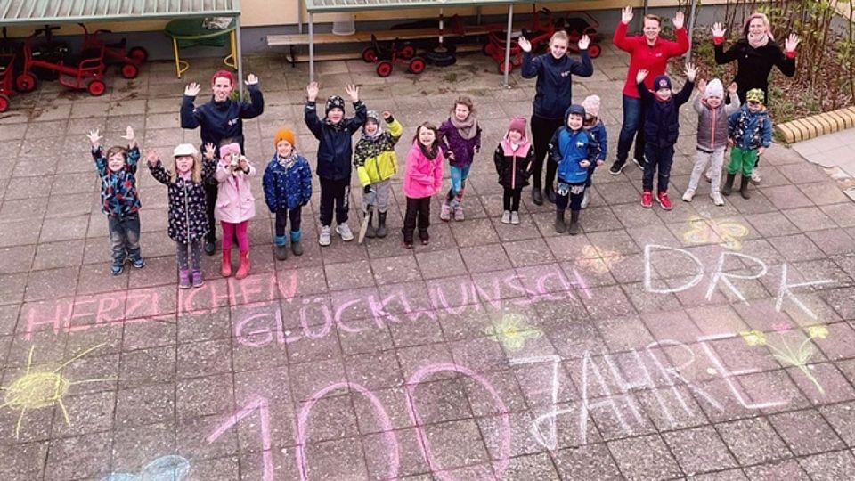 5fd8b8f4-3c49-47dc-a3c1-f7519a634edc Bild zeigt Kita-Kinder mit Erziehern auf dem Innenhof der Kita. Alle winken mit den Händen und schauen nach oben da das Bild von oben aufgenommen wurde. Auf dem Boden steht mit farbiger Kreide der Schriftzug Herzlichen Glückwunsch 100 Jahre DRK