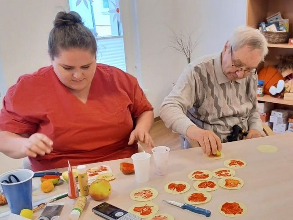 Bild zeigt Frau Holtz und Herrn Wendel an einem Tisch sitzend. Vor ihnen liegen Apflehälften, rote und grüne Farbe sowie Pinsel und Becher mit Wasser. Herr Wendel hält eine mit roter Farbe bestrichene Apfelhälfte über ein rundes Stück Papier um im nächsten Augenblick den Apfel auf das Papier zu pressen. Vor ihm liegen schon etliche runde Blätter mit dem aufgedruckten Muster der Apfelhälften.