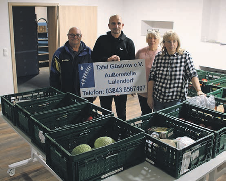 Bild zeigt die in der Bildunterschrift genannten 4 Personen von links nach rechts. Herr Marzahn hält das Schild in den Händen . Auf den Tischen vor den Personen stehen grüne Kisten mit Lebensmitteln die zur Verteilung vorgesehen sind.