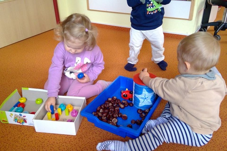 Bild zeigt: Drei Kleinkinder spielen gemeinsam auf dem orangefarbenen Boden eines Gruppenraums mit verschiedenen Lernmaterialien.  Ein Mädchen in einem lila Pullover kniet und sortiert bunte Steckspiele in einer weißen Kiste.  Ein weiteres Kind, in einem beigefarbenen Oberteil und blau-weiß gestreiften Hosen, sitzt und spielt mit Kastanien und einem roten Steckdosenbehälter in einer blauen Plastikschale.  Im Hintergrund steht ein drittes Kind.  Die Szene zeigt eine Freispielsituation im Kindergarten, in der die Kinder durch vielfältige Materialien zum Entdecken und Lernen angeregt werden.