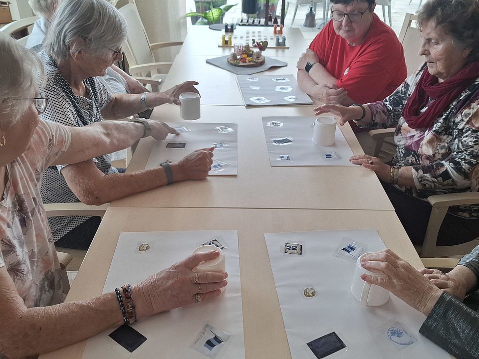 Bild zeigt mehrere Tagesgäste sitzend an einem langen Holztisch in einem hellen Raum und nehmen an einem Konzentrationsspiel teil. Vor jeder Person liegen zwei weiße Blätter mit jeweils mehreren   aufgedruckten Symbolen. Die Seniorinnen halten weiße Becher in der Hand, die sie über die Symbolbilder bewegen, um das angesagte Symbolbild zu verdecken. Eine Betreuerin in einem roten DRK T-Shirt sitzt am Ende des Tisches und beobachtet das Geschehen.