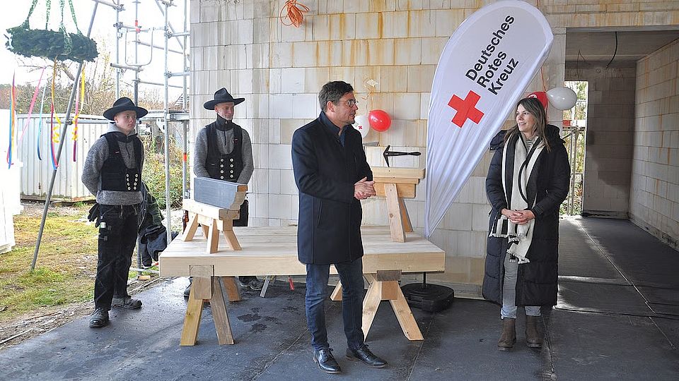 Bild 1 Bild zeigt zeigt den Landrat Sebastian Constin auf der Baustelle in einem überdachten Bereich. Neben ihm steht Frau Carolin Schwarz vom DRK Güstrow. Herr Constin hält eine Rede. Im Hintergrund sind zwei Zimmermannsleute in Ihrer Tracht-Arbeitskleidung zu sehen. Weiter im Hintergrund sieht man den Richtkranz am Gebäude hängen.