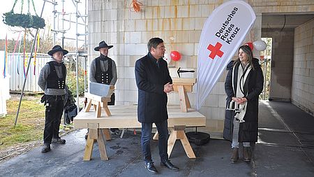Bild zeigt zeigt den Landrat Sebastian Constin auf der Baustelle in einem überdachten Bereich. Neben ihm steht Frau Carolin Schwarz vom DRK Güstrow. Herr Constin hält eine Rede. Im Hintergrund sind zwei Zimmermannsleute in Ihrer Tracht-Arbeitskleidung zu sehen. Weiter im Hintergrund sieht man den Richtkranz am Gebäude hängen.