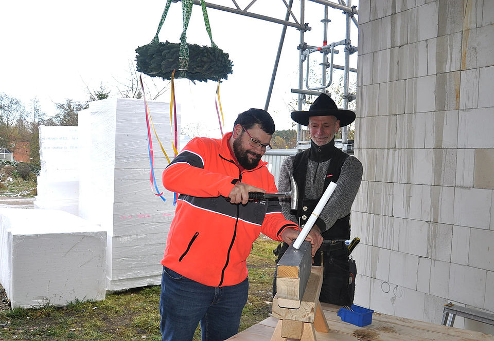 Bild 2 Bild zeigt Herrn Henneberg von der Sozialstation Krakow am See mit einem Zimmermannshammer vor einem Balken, in den er versucht den "letzten Nagel" einzuschlagen. Herr Asse in seiner Zimmermannstracht hält den Balken mit fest und schaut genau hin, damit nichts schief geht. Beide Personen haben lächelnde Gesichter.