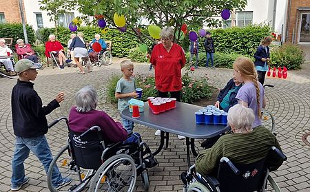 Bild zeigt einige Kinder und Senioren im Rollstuhl bei einem Ping-Pong-Becher Spiel an einem Tisch der im Freien aufgestellt ist