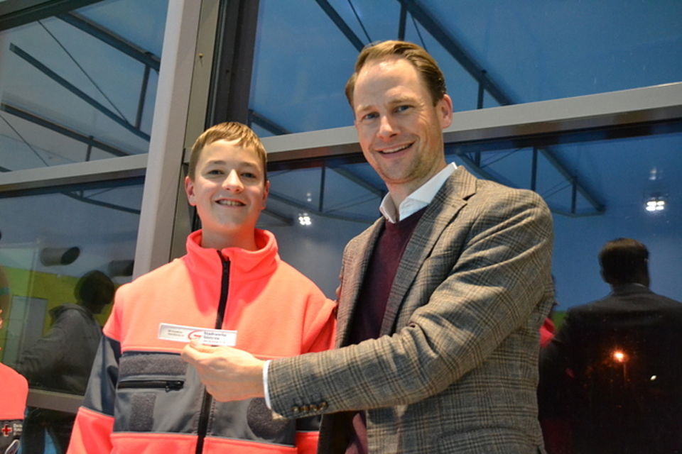 Juniorretter Bild 3 Bild zeigt Herrn Graßhoff neben einem Jungen, Hans-Erik, in der Hand hält Herr Graßhoff den Sponsoring-Patch, welcher dann gleich nach dem Foto auf Hans-Erik's DRK Jacke angebracht wird.