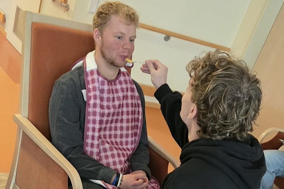 Bild zeigt zwei junge Männer, einer sitz auf einem Lehnenstuhl halt ein Latztuch um. der andere junge Mann reicht dem auf dem Stuhl sitzenden einen Löffel mit Brei an den Mund.