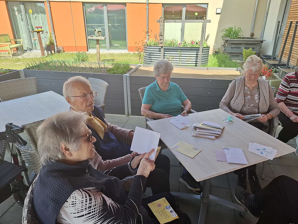 Bild zeigt vier Tagesgäste an einem Tisch sitzend. Auf dem Tisch liegen viele Brief in Umschlägen, einige sind schon ausgepackt. Ein Frau vorne links am Tisch liest eine Karte, die sie in ihren Händen hält, laut vor.